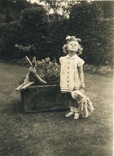 A little girl stands to attention and grimaces for the camera in a garden holding a doll