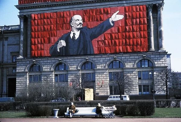 A banner of lenin on the side of a building overlooking two elderly women sitting on a park bench in leningrad, ussr, 1960s