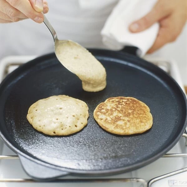 Blinis cooking on frying pan, raw mixture being added with a spoon, high angle view