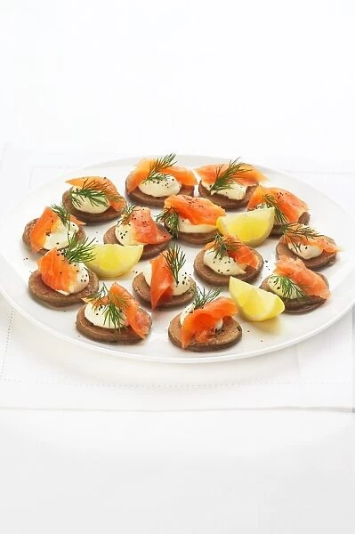 Blinis topped with smoked salmon, creme fraiche, dill and black pepper and garnished with lemon wedges, on a plate, high angle view