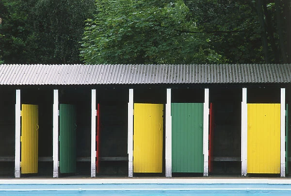 Changing rooms with brightly painted doors and corrugated iron rook, standing by an outdoor swimming pool