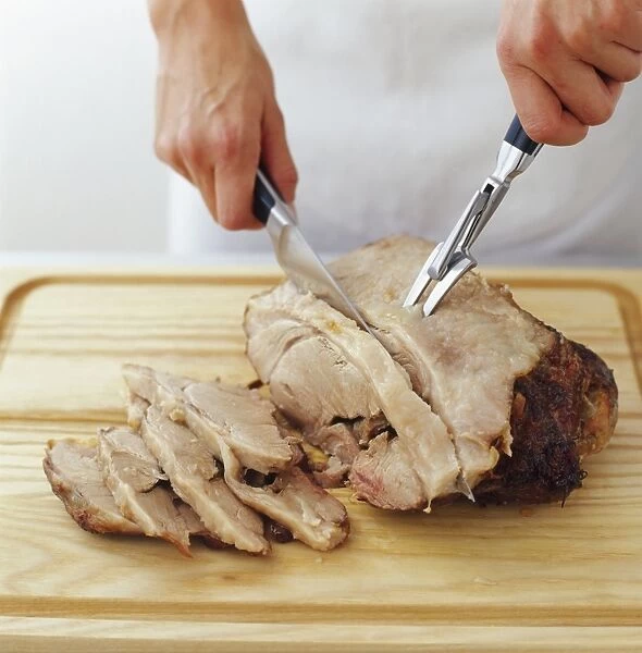 Chef using carving knife and fork to cut roast shoulder of pork into thick slices on wooden chopping board