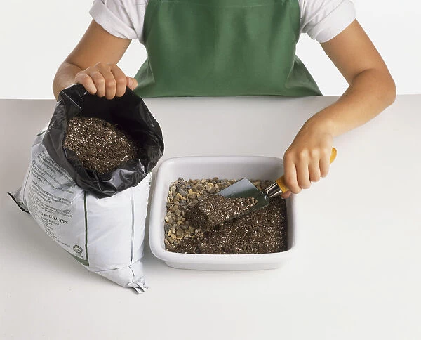 Child preparing white plant container; covering layer of gravel with potting compost, using trowel, steading compost bag beside