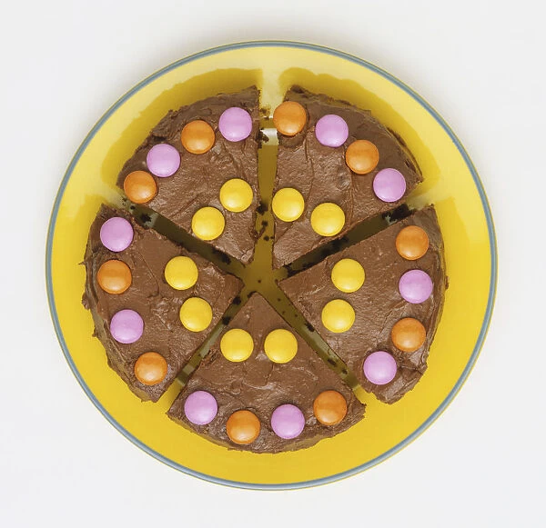 Chocolate cake decorated with colourful sugar-coated confectionery, cut into five pieces, served on yellow plate, view from above