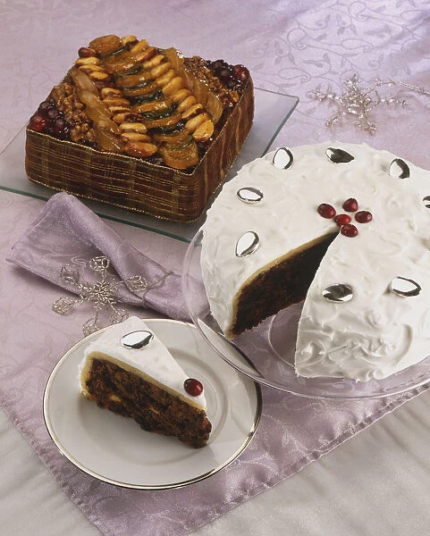 Two Christmas fruit cakes on festive tablecloth, whole square candied cake, iced circular cake with removed slice on serving plate, elevated view