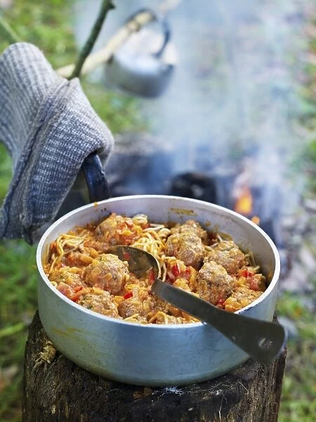Cooked meatballs and pasta sauce in aluminium pan on top of tree stump near camp fire