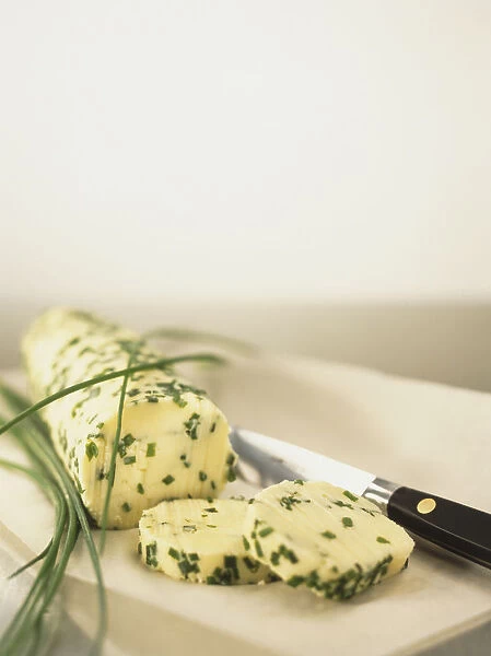 Cylindrical block of herb butter with chives, served on rectangular tray with knife, close up