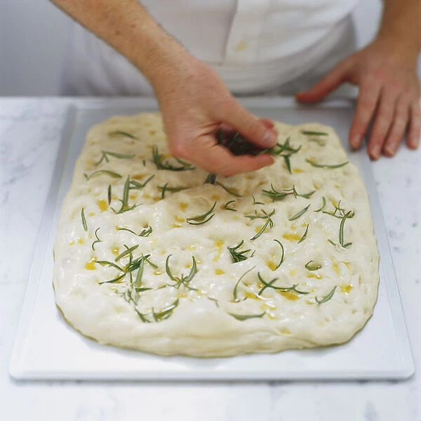 Fresh rosemary sprigs being pressed into uncooked focaccia dough, high angle view