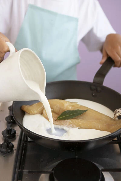 Girl pouring milk into frying pan containing piece of haddock and bay leaf