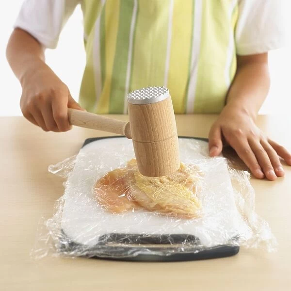 Girls hand using mallet to tenderise chicken breasts, covered with clingfilm, close-up