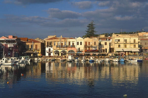 Greece, Greek Islands, Crete, Rethymno, harbor lined with shops and boats moored at harborside, their hulls and rigging reflected in water