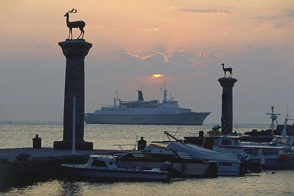 Greece, Rhodes, Mandraki harbour with two statues of deer at its entrance, at sunset, ferry in background