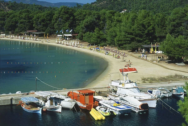Greece, Skiathos, sweeping bay of Koukounaries, with sunbathers, and boats moored in a small harbour area, wooded area behind