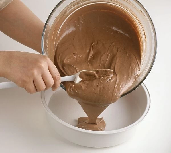 Hand holding mixing bowl and spoon, pouring chocolate cake mixture into cake tin