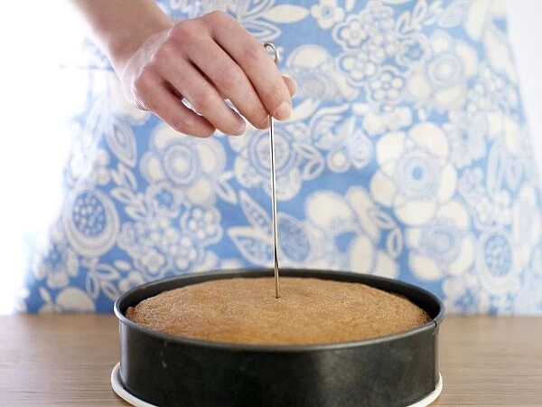 Hand inserting metal skewer into baked cake in round cake tin, close-up