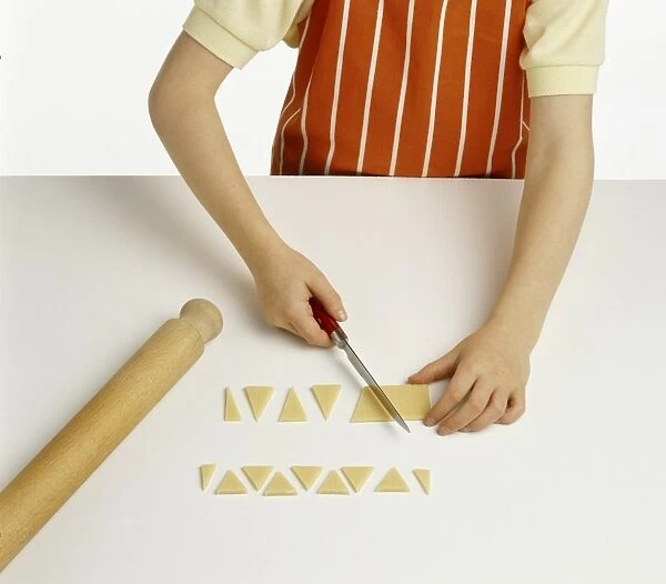 Hand model wearing orange and white striped apron, cutting triangular shapes of plain marzipan, rolling pin beside