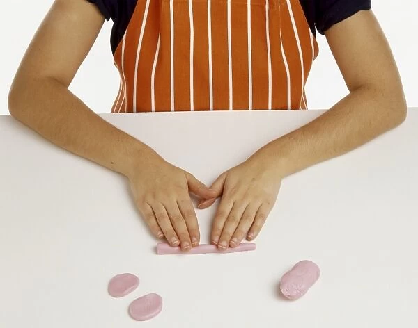 Hand model wearing an orange and white striped apron, making oval shapes from pink fondant icing, rolling icing into long bar