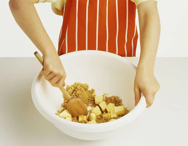 Hand model wearing an orange and white striped apron, mixing together butter and soft brown sugar in a large white mixing bowl