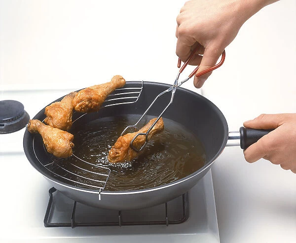 Hands using tongs to move deep-fried chicken drumstick from pan onto draining rack
