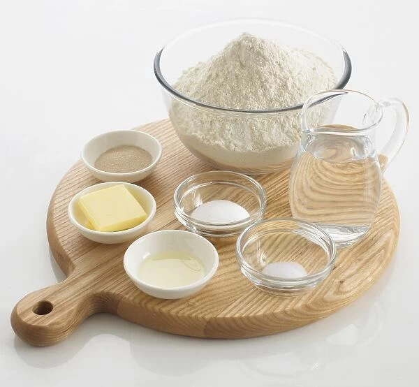Ingredients required to make bread, including water, flour, salt, sugar, butter, yeast, in bowls and a jug, arranged on a chopping board