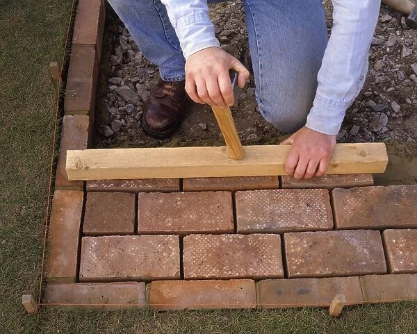 Laying brick paving, tamping the bricks level using a piece of wood and the reverse end of a hammer, close-up
