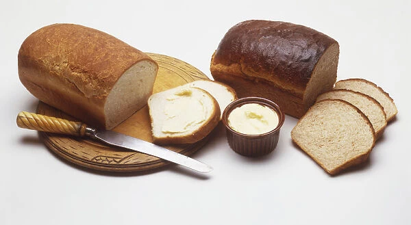 Loaves of white and brown bread, some slices cut away, a bowl of butter, a knife and a chopping board