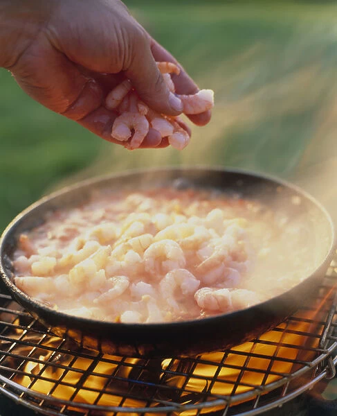 Man adding prawns to salsa fresca cooking in heavy pan over barbecue grill