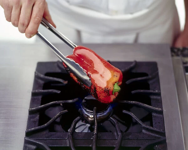 Man charring a red pepper by holding over an open flame using long-handled kitchen tongs
