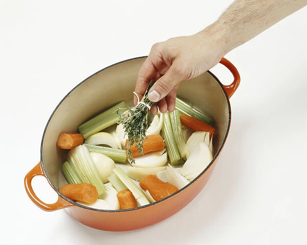 Man holding bouquet garni above onions, carrots and celery in casserole dish in preparation for making vegetable stock