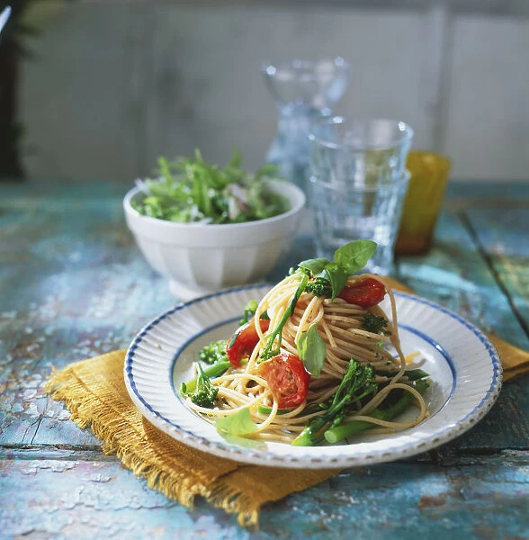 Plate of spaghetti with broccoli, cherry tomatoes and garnish leaves, bowl of green salad and stacked empty glasses in background, front view