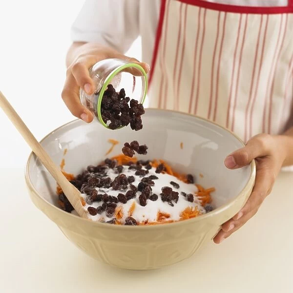 Pouring raisins from bowl onto carrot cake mixture in mixing bowl, close-up