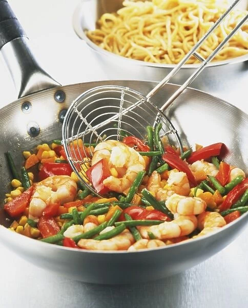 Prawn and vegetable stir-fry in a wok with wire spoon, bowl of noodles in background
