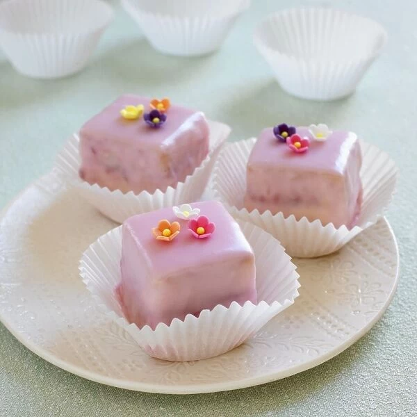 Raspberry cakes with pink icing and flower decorations on a plate, cake cases in the background