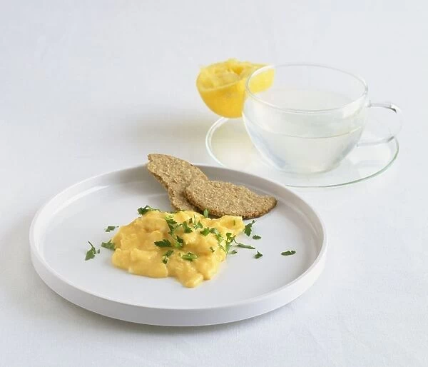 Scrambled eggs with parsley and oatcake on white plate next to cup of hot water and lemon