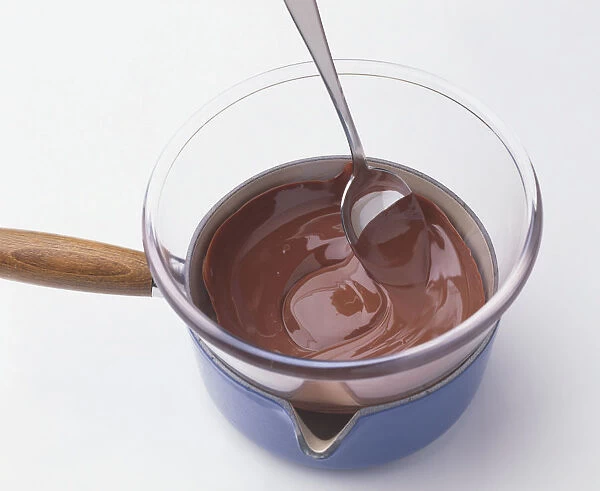 Spoon in melted chocolate in glass mixing bowl on top of saucepan containing boiling water, close-up