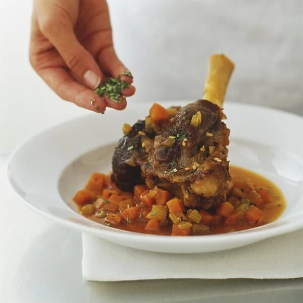 Sprinkling chopped rosemary over a dish of braised lamb shanks, served with a sauce of diced carrots and celery