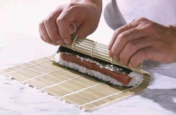 Sushi containing nori seaweed, rice and tuna, being hand-rolled inside bamboo mat, close-up