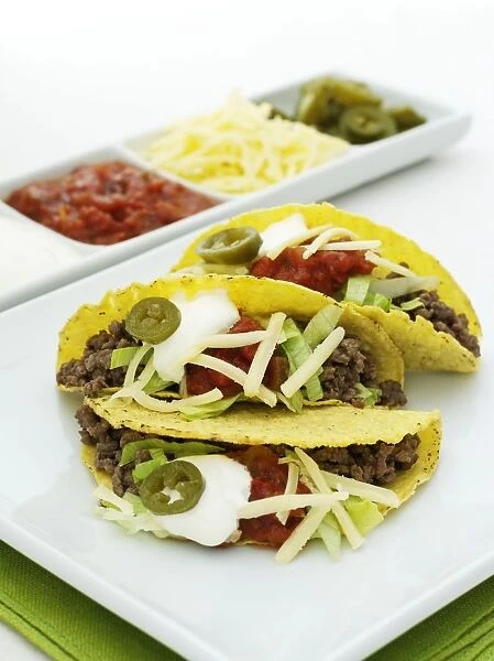Tacos stuffed with minced beef, lettuce, cheddar cheese, tomato salsa, sour cream and jalapeno peppers, tray containing salsa, cheese and peppers in the background
