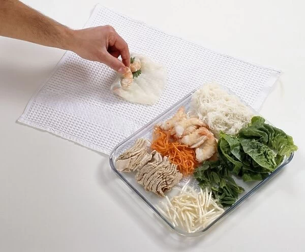 Using fingers to roll prawns in rice paper on cloth next to ingredients for Vietnamese spring rolls on chopping board