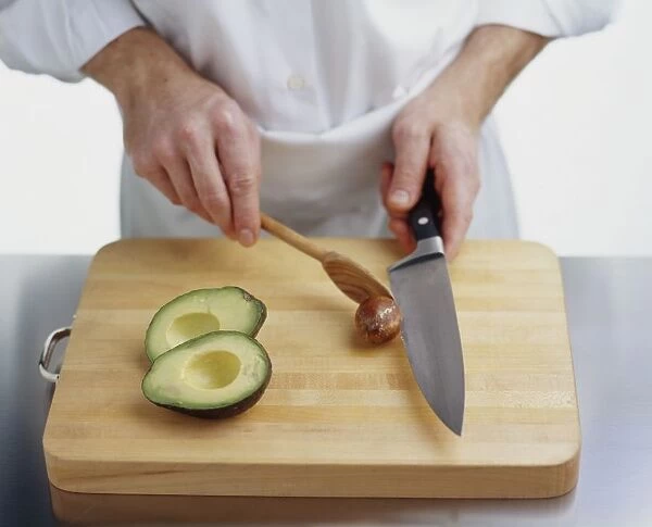 Using wooden spoon to pry avocado stone off knife blade, two avocado halves on chopping board