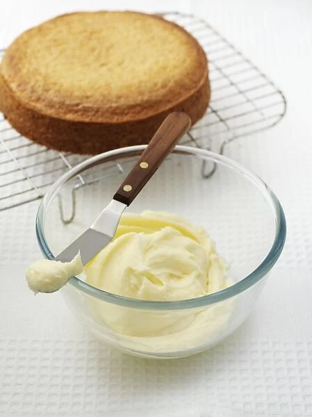 Vanilla buttercream icing in glass bowl and on cake spatula, cake on cooling rack nearby, close-up