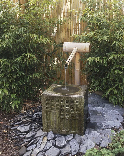 Water trickling through bamboo pipe and spout into square basin, in front of bamboo garden fence