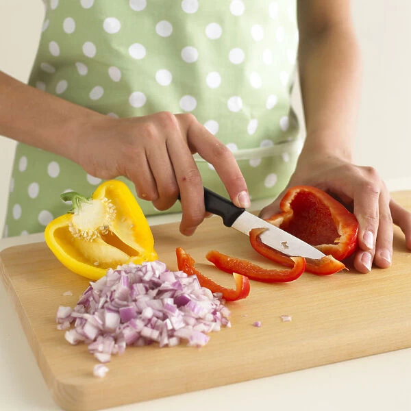 Woman slicing red pepper next to diced red onion and half yellow pepper