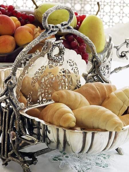 Antique silver hinged bread basket with croissants in front of a bowl of fruit on a breakfast table
