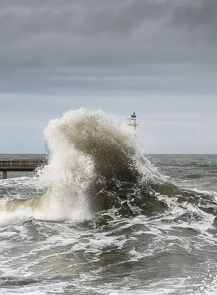 Large Wave Crashing At The Coast With A Lighthouse At The End Of A Pier; Amble, Northumberland, England