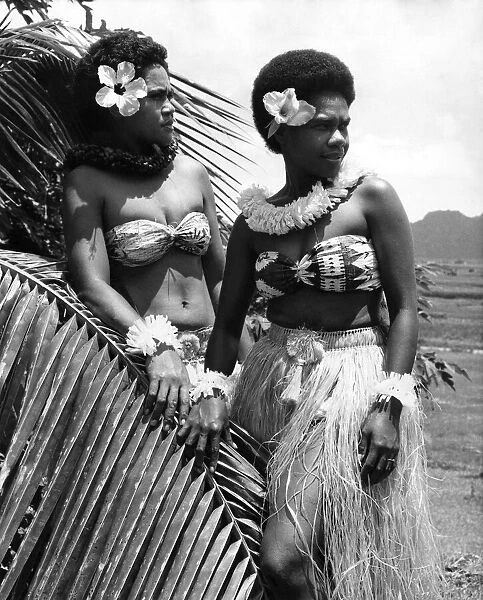 Fiji: Two girls in grass skirts against a background of palms, flowers and mountains