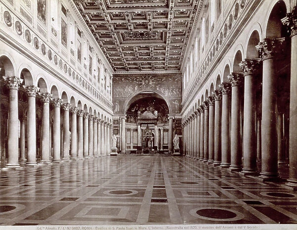 The nave of the Basilica of Saint Paul Outside the Walls, in Rome. A colonnade of smooth columns with Corinthian capitals separates the nave from the aisles. A frieze with mosaic portraits of the Popes in tondos runs along above the walls. In the upper register, between the windows, are frescoes with scenes from the life of Saint Paul. In the background, the statues of Saint Peter by Jacometti and of Saint Paul by Ravelli. The majestic Triumphal Arch has mosaics taken from the original arch dating to the time of Saint Leo the Great. The ciborium over the high altar is by Arnolfo di Cambio, the apse mosaic dates to Honorius III. The coffered ceiling is a copy in Sixteenth century style of the one destroyed in the fire of 1823. The marble floor has geometric patterns
