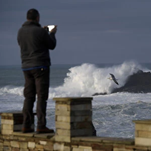 A man takes pictures of waves as they crash against a lighthouse in the port town of