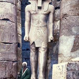 EGYPT: LUXOR TEMPLE. A man seated next to a statue of Pharaoh Ramesses II at the Luxor Temple