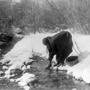 Apsaroke woman standing in snow scooping water from a stream with a can, bucket beside her